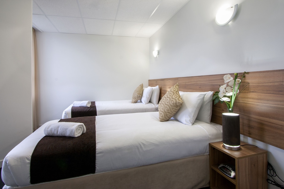 A hotel room with two beds, each with white and brown bedding, a wooden headboard, and a bedside table with a lamp.