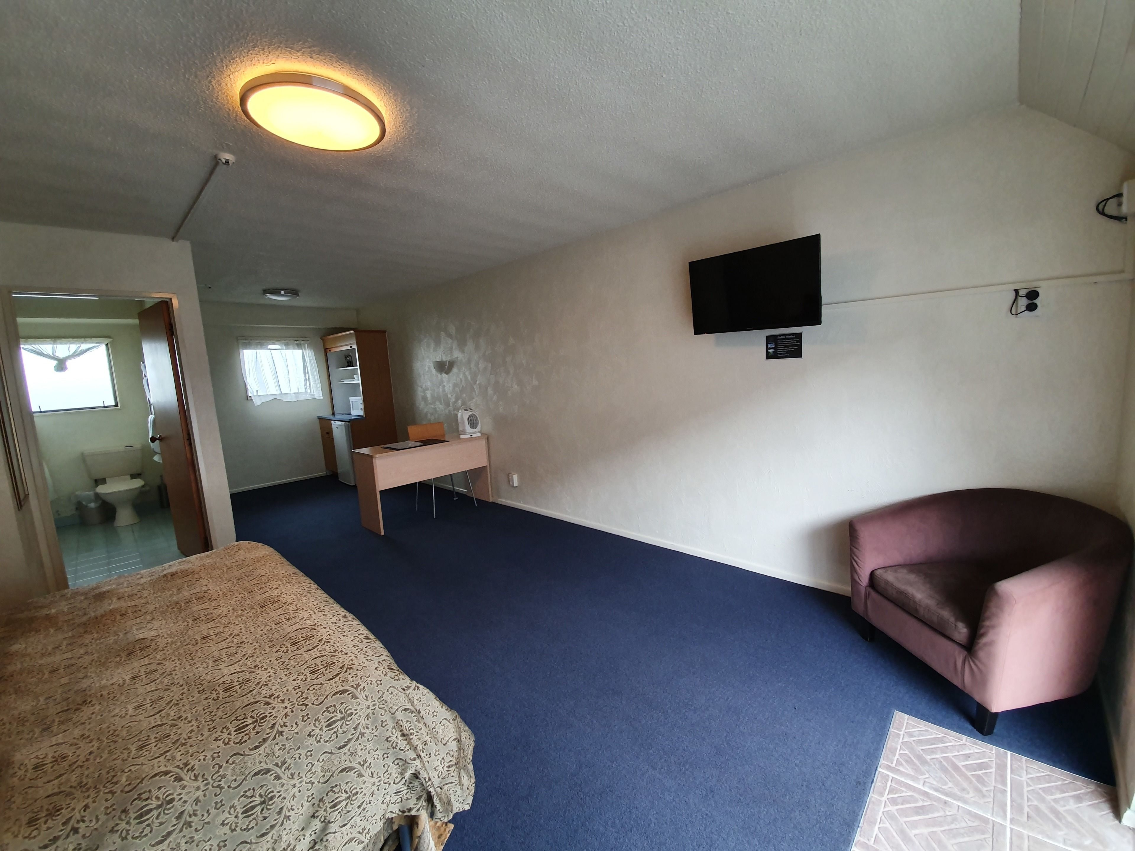 Ein Schlafzimmer mit Bett, Schreibtisch, Stuhl, Fernseher und einem durchgehenden Türspalte sichtbaren Badezimmer.