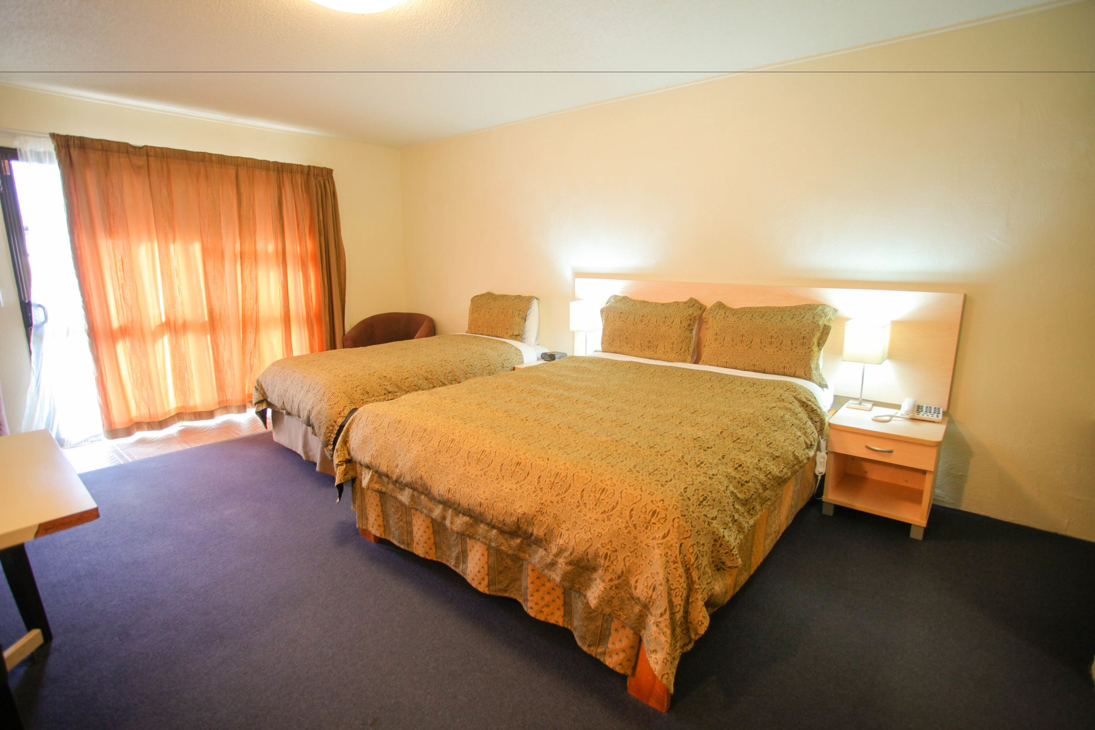 Quarto de hotel com duas camas, uma escrivaninha, uma cadeira e uma mesa de cabeceira com abajur.