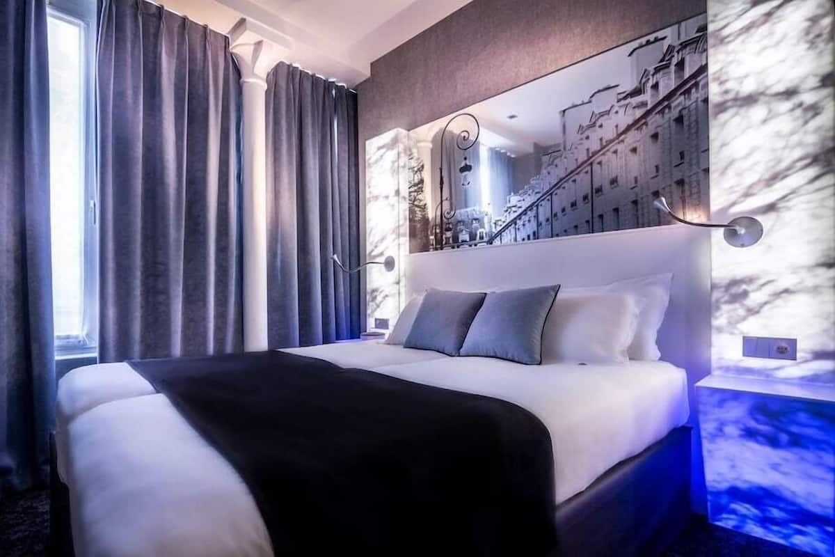 A modern hotel room with a large bed, grey curtains, a cityscape mural, and a staircase.