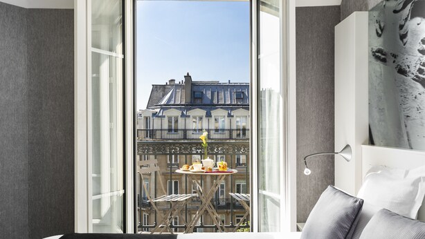 A hotel room with a balcony, a dining table with a vase of flowers, and a bed with pillows. - Hotel Marais Grands Boulevards (Paris)