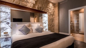 A modern bedroom with a stone wall, a large bed with white bedding and two pillows, a black throw blanket, a bedside table with a lamp, and a glass shower door. - Hotel Marais Grands Boulevards (Paris)