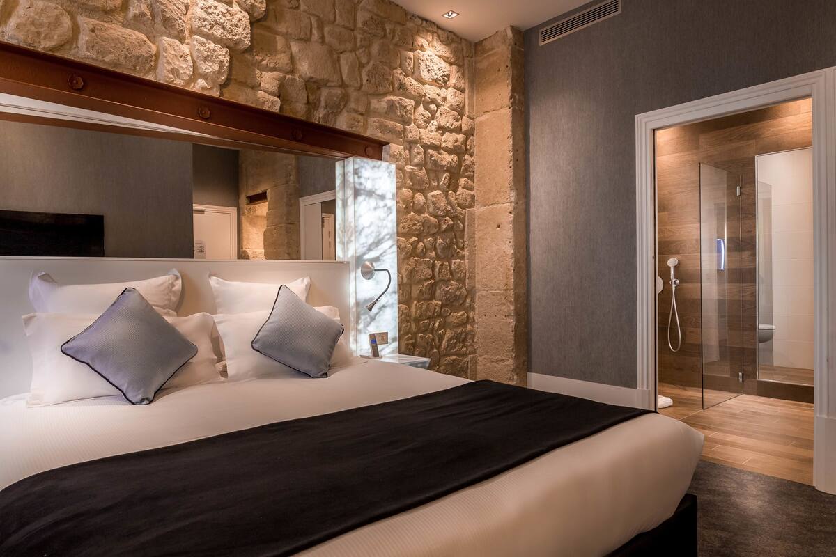A modern bedroom with a stone wall, a large bed with white bedding and two pillows, a black throw blanket, a bedside table with a lamp, and a glass shower door.