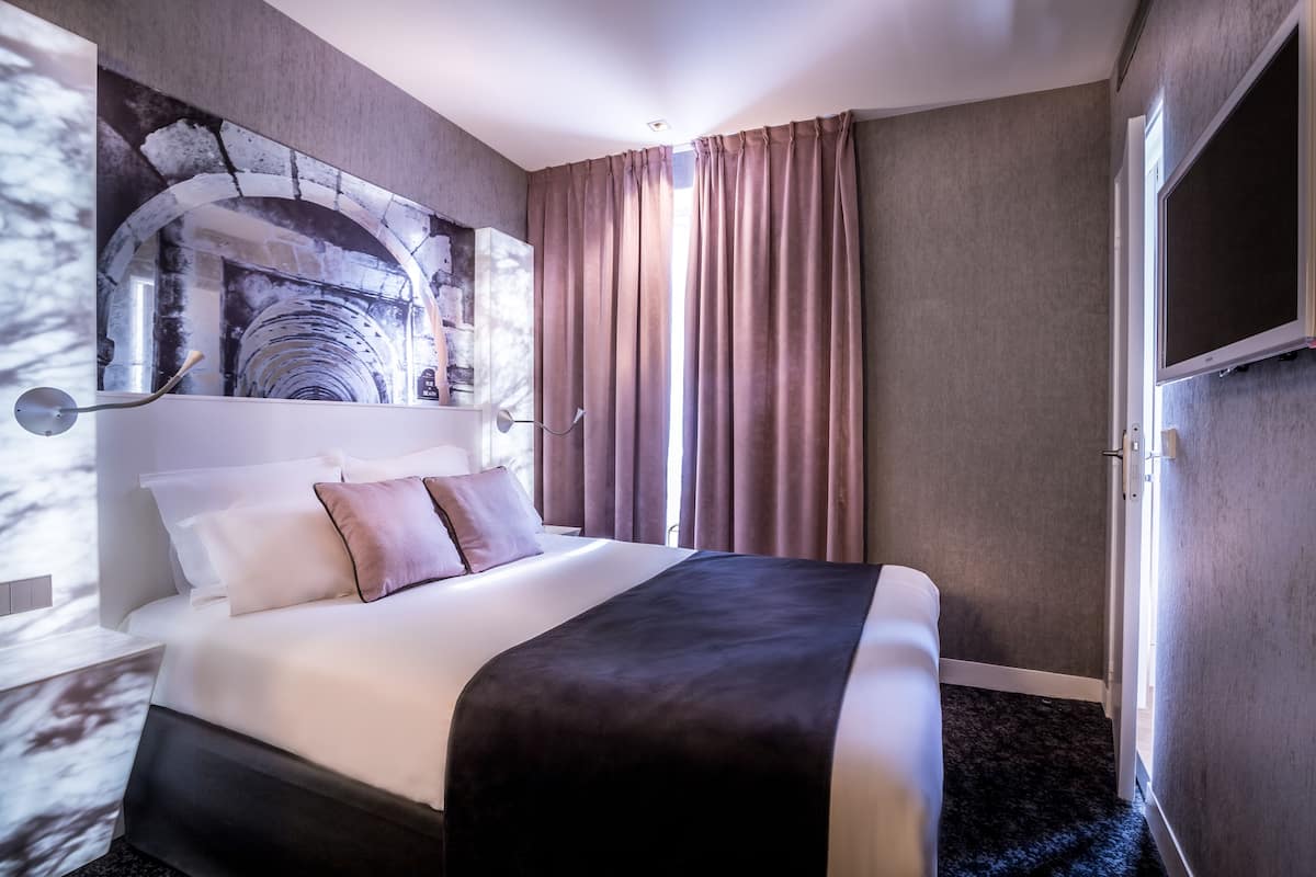 A hotel room with a large bed, a stone arch wall art, a flat-screen TV, and a window with curtains.
