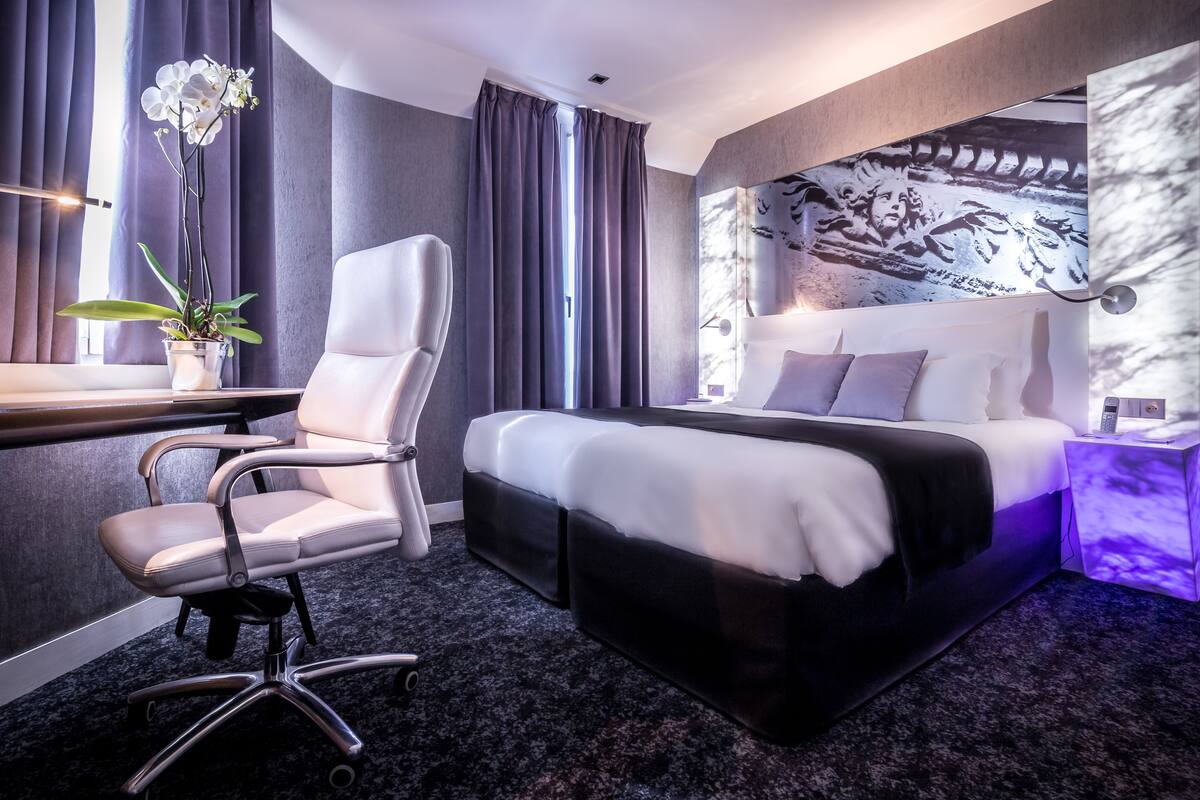 A modern hotel room with a large bed, a desk with a chair, a potted plant, and a wall-mounted artwork.