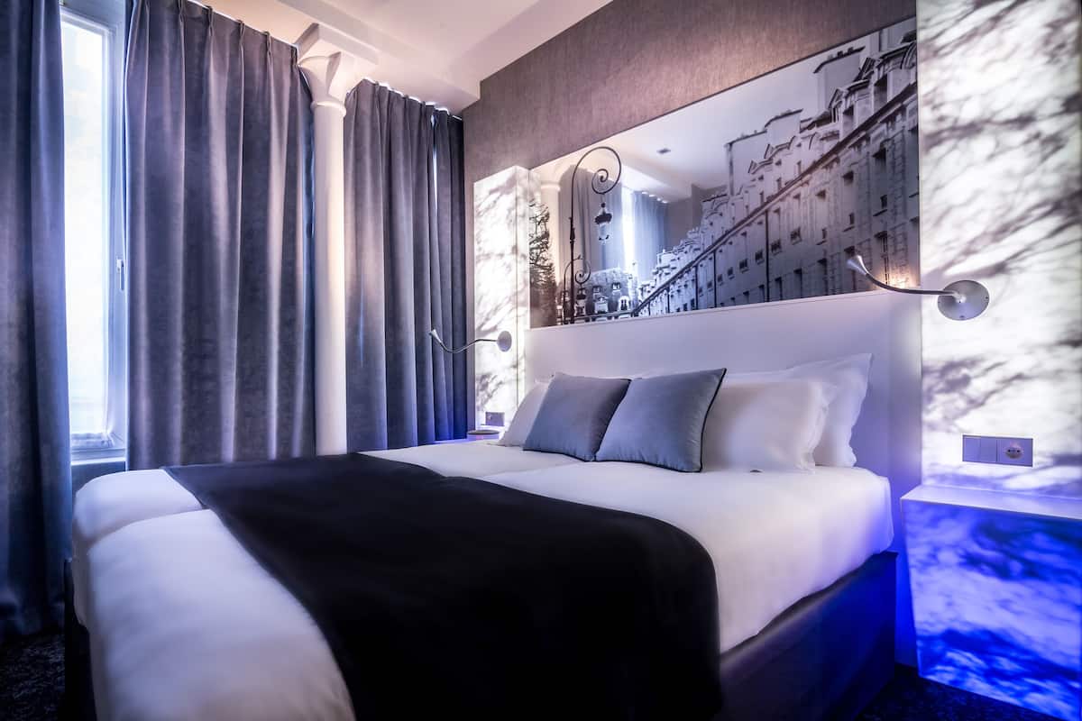 A modern hotel room with a large bed, grey curtains, a cityscape mural, and a nightstand with a lamp.
