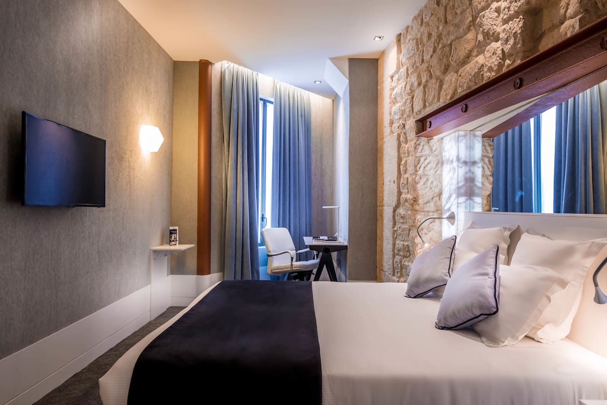 A modern hotel room with a large bed, a flat-screen TV mounted on the wall, a desk with a chair, and a stone accent wall.