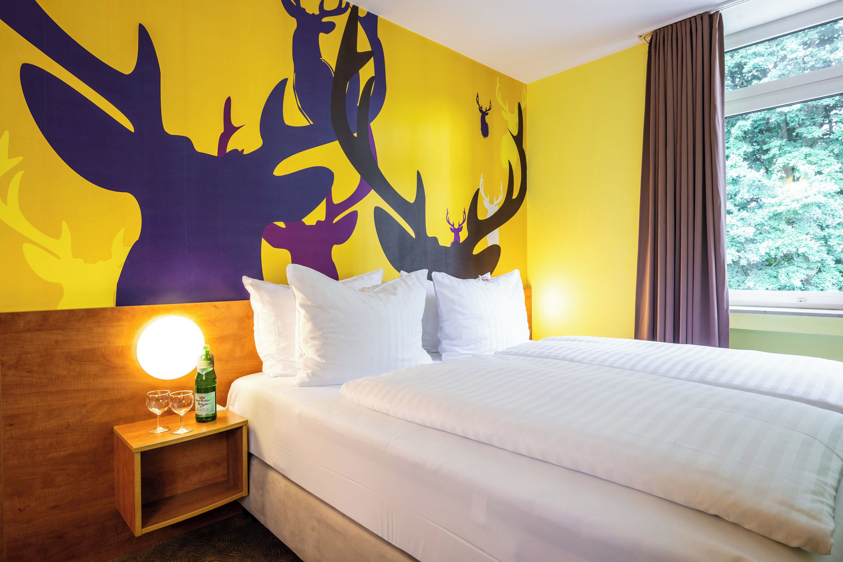 Ein Hotelzimmer mit Bett, weißen Kissen, einem Nachttisch mit Flasche und Gläsern, Wandgemälde von Hirschen und einem Fenster mit Vorhängen.