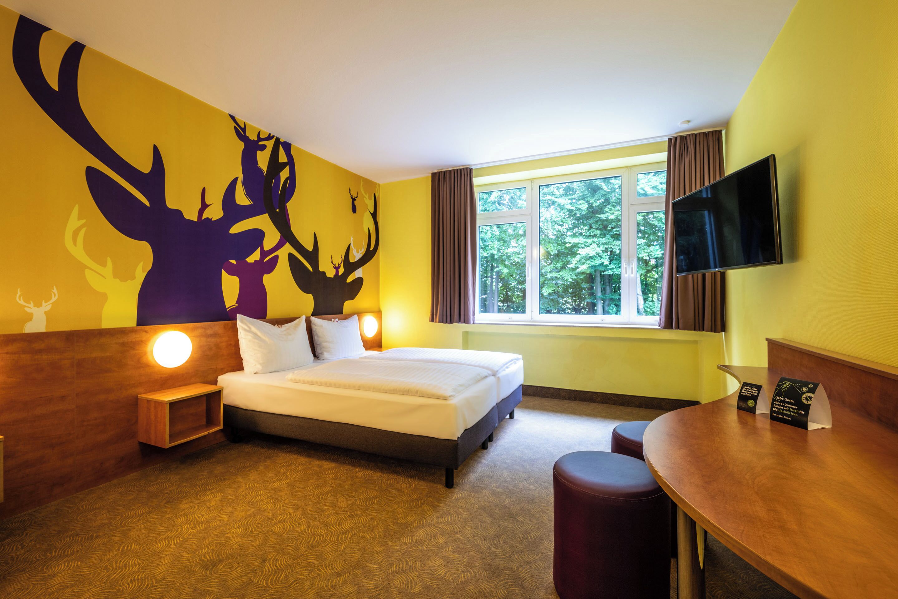 Ein Hotelzimmer mit Bett, Schreibtisch, Fernseher, einem Fenster mit Vorhängen und einem Wandbild von Hirschen.