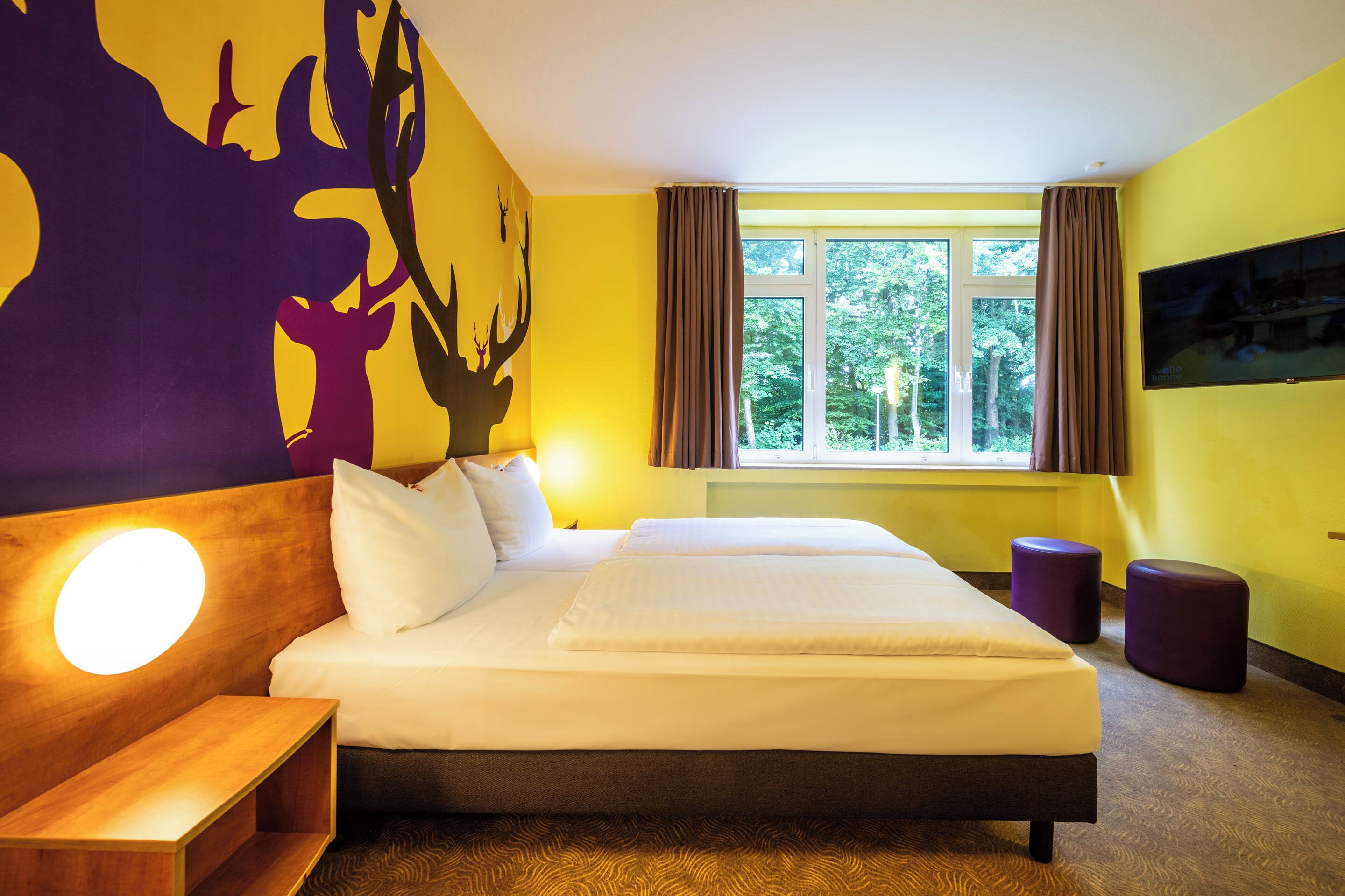 Habitación de hotel con una cama grande, una mesita de noche, televisor montado en la pared y una ventana con cortinas.