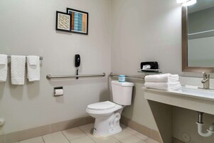 Un baño con inodoro blanco, barra de apoyo, lavabo con espejo y toallero con toallas dobladas.