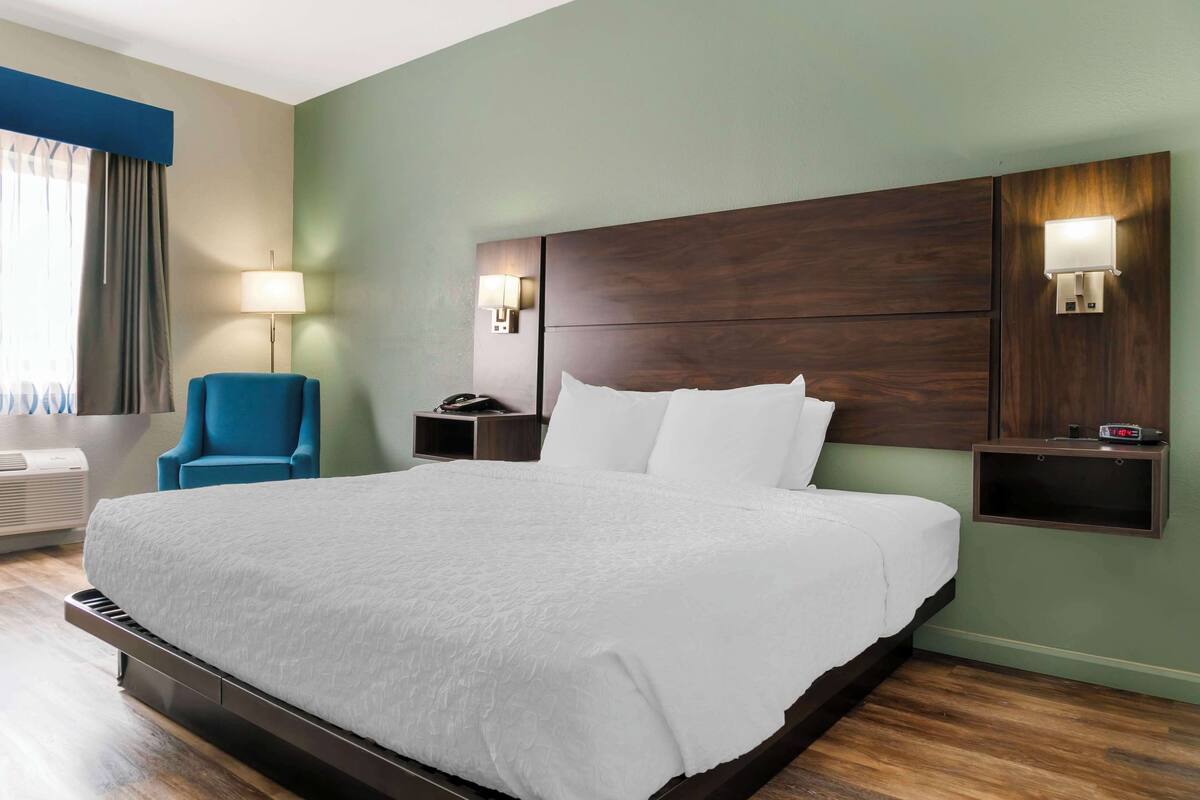 A hotel room with a large bed, a blue armchair, bedside lamps, and a small table with a clock.