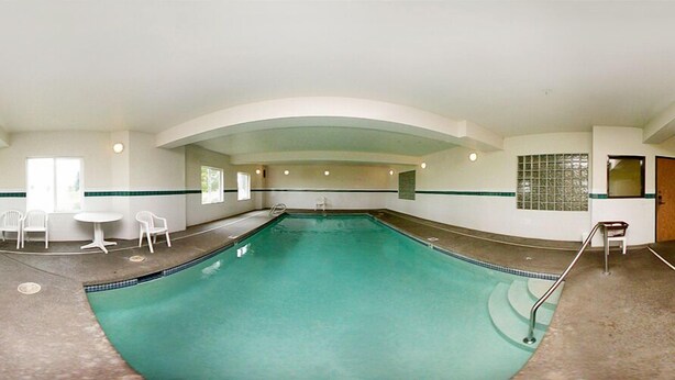 Una grande piscina interna dalla forma sinuosa, circondata da pareti bianche e da una fascia verde. Vicino alla piscina, ci sono sedie bianche e un piccolo tavolo.