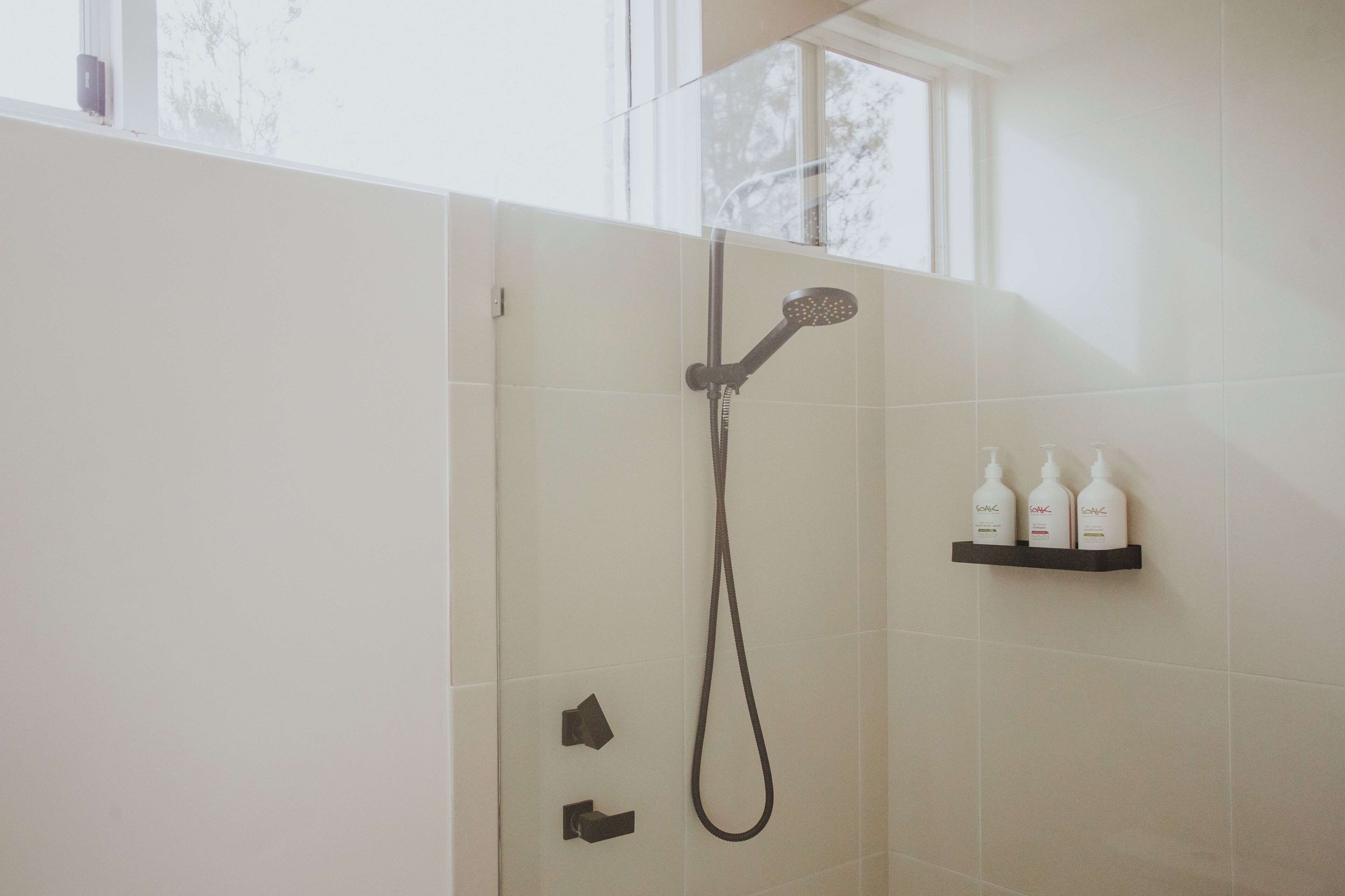 A shower with a handheld showerhead and a wall-mounted faucet. There are three bottles of shampoo on a shelf above the shower.