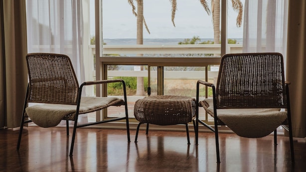 Two wicker chairs with cushions, positioned in front of a large window with a view of palm trees and the ocean. - The Ellson Hotel (Kingscote)