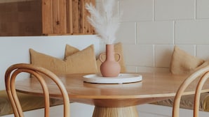 A wooden dining table with a pink vase and white feathers, surrounded by wooden chairs. - The Ellson Hotel (Kingscote)