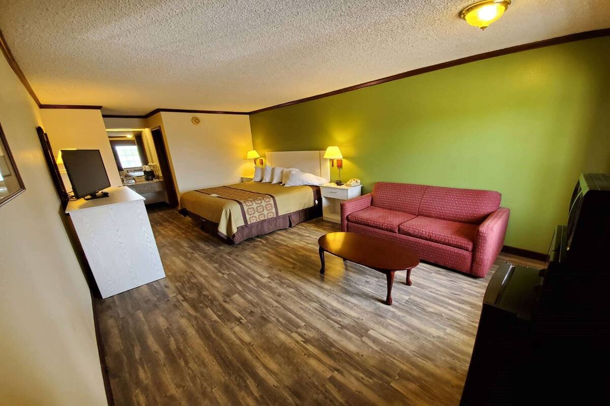 A hotel room with a bed, a red sofa, a small table, a desk with a lamp, and a television.