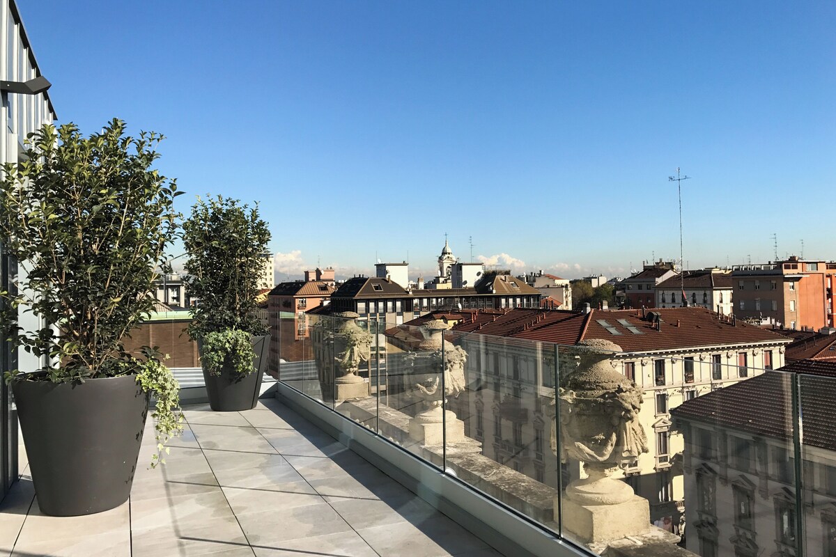 A rooftop terrace with potted plants, a glass railing, and a view of surrounding buildings.