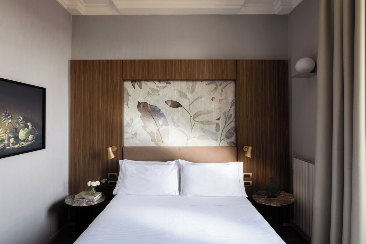 A hotel room with a bed, a headboard with a leaf pattern, two bedside tables with lamps, and a framed picture on the wall.