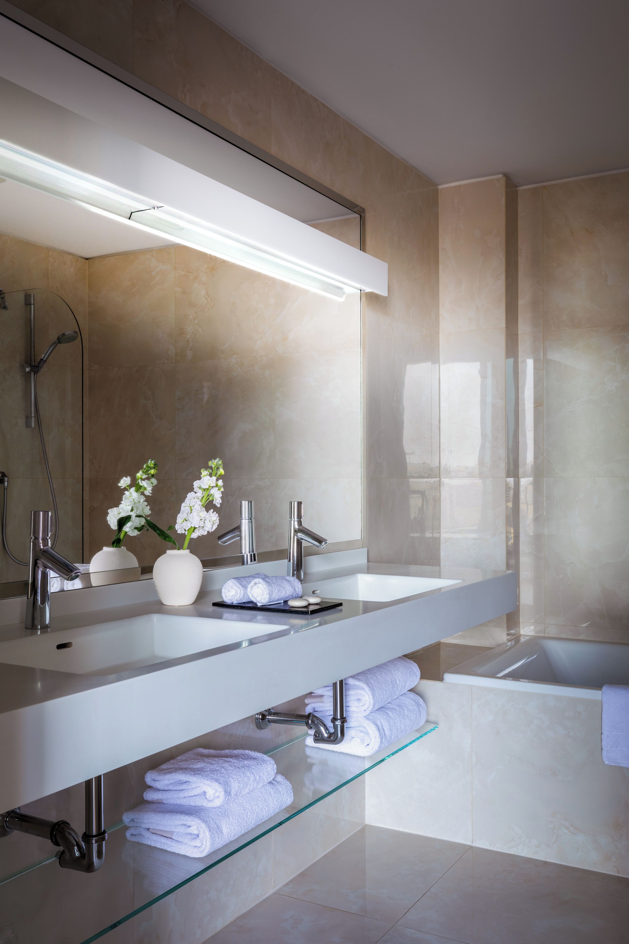 A modern bathroom with two sinks, a large mirror, a glass shelf with towels, and a bathtub.