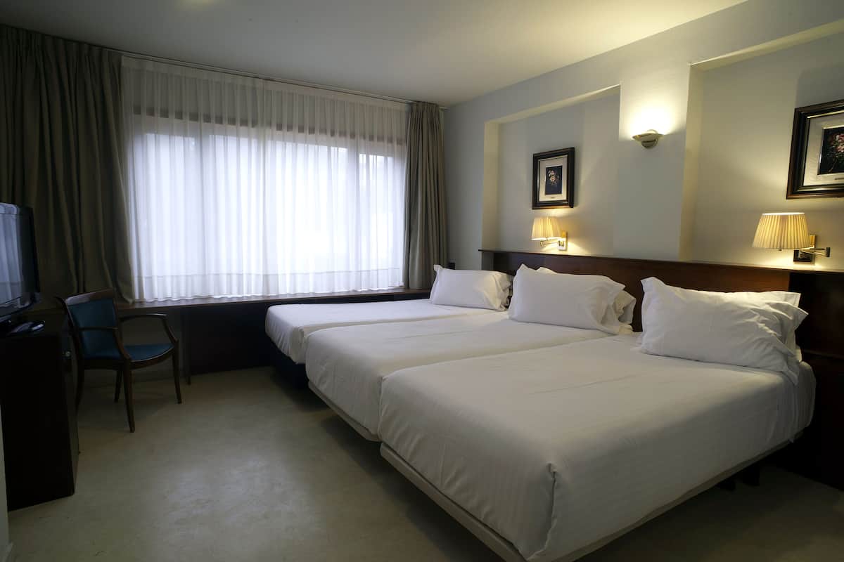 A hotel room with two beds, a desk, a chair, and a large window with curtains.
