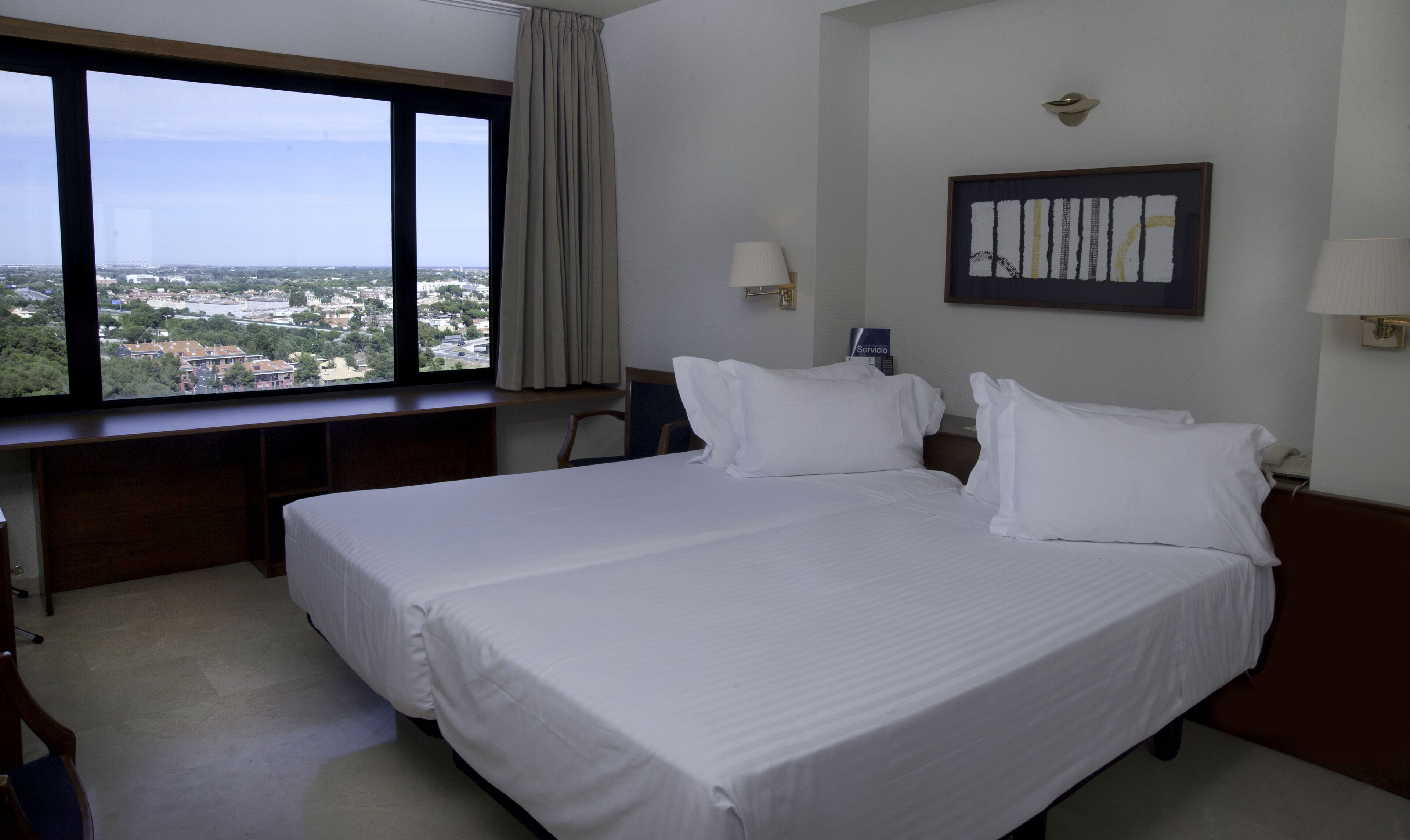 A hotel room with a large bed, a desk, and a view of the city through a large window.