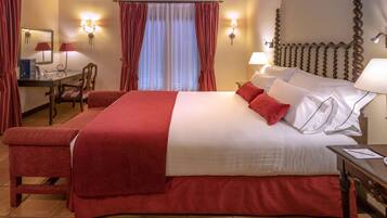 A hotel room with a large bed, red curtains, a desk with a chair, and wall-mounted lamps.
