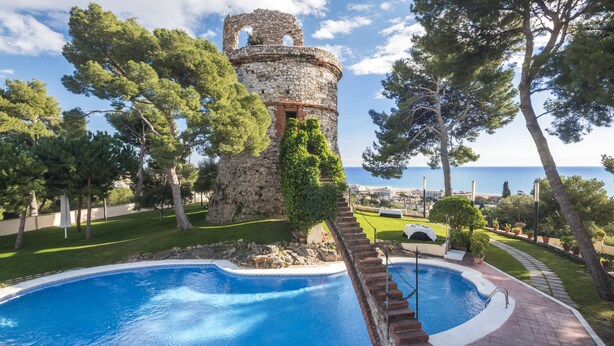 A swimming pool with a stone tower, surrounded by trees and a view of the sea. - Gran Hotel Rey Don Jaime (Castelldefels)