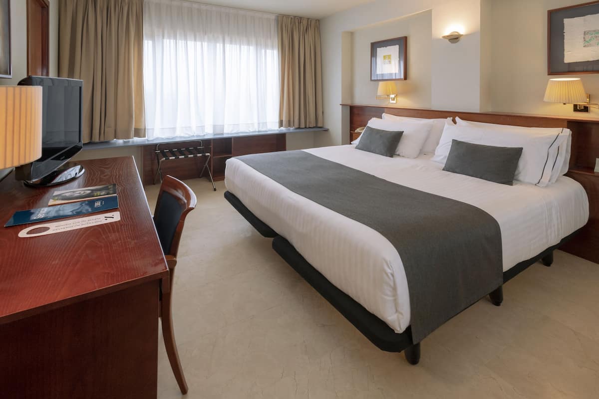 A hotel room with a bed, desk, chair, and a window with curtains.