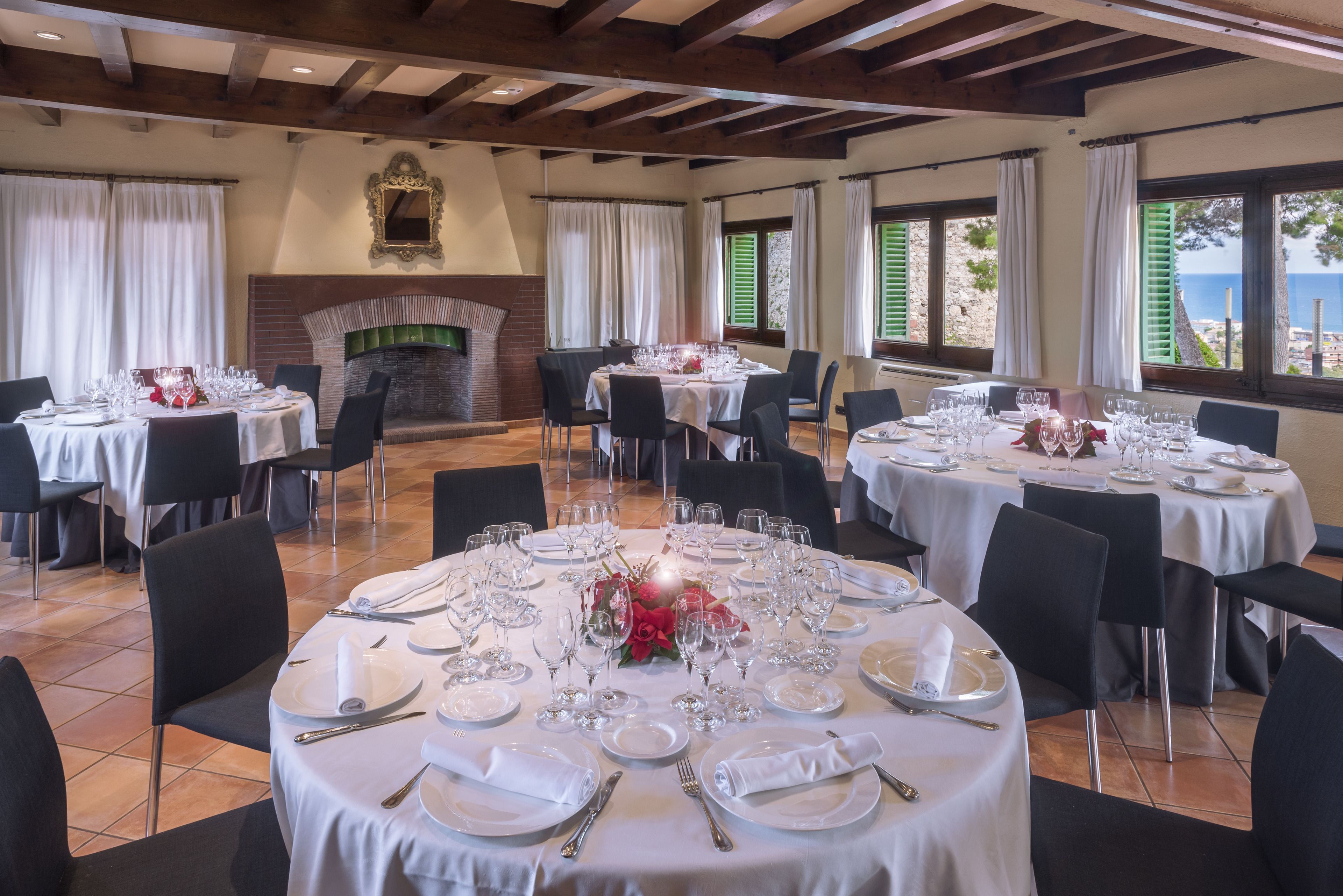 A dining area with round tables set for a meal, a fireplace, and a view of the sea.