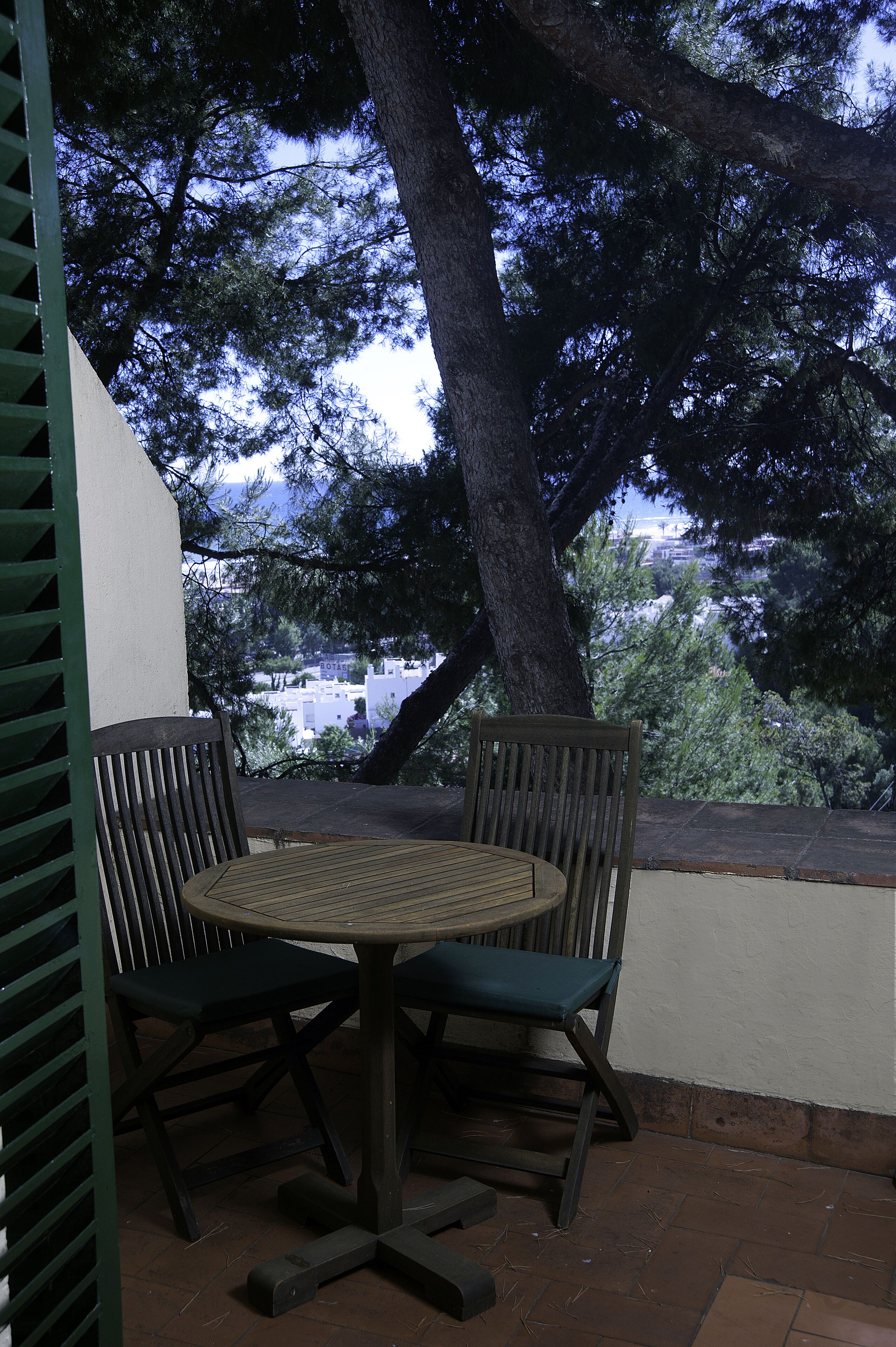 A patio with a round table and two chairs, surrounded by a large tree and a view of buildings in the background.