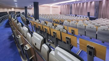 Un amplio salón de conferencias con filas de sillas, un escenario y un telón de fondo.