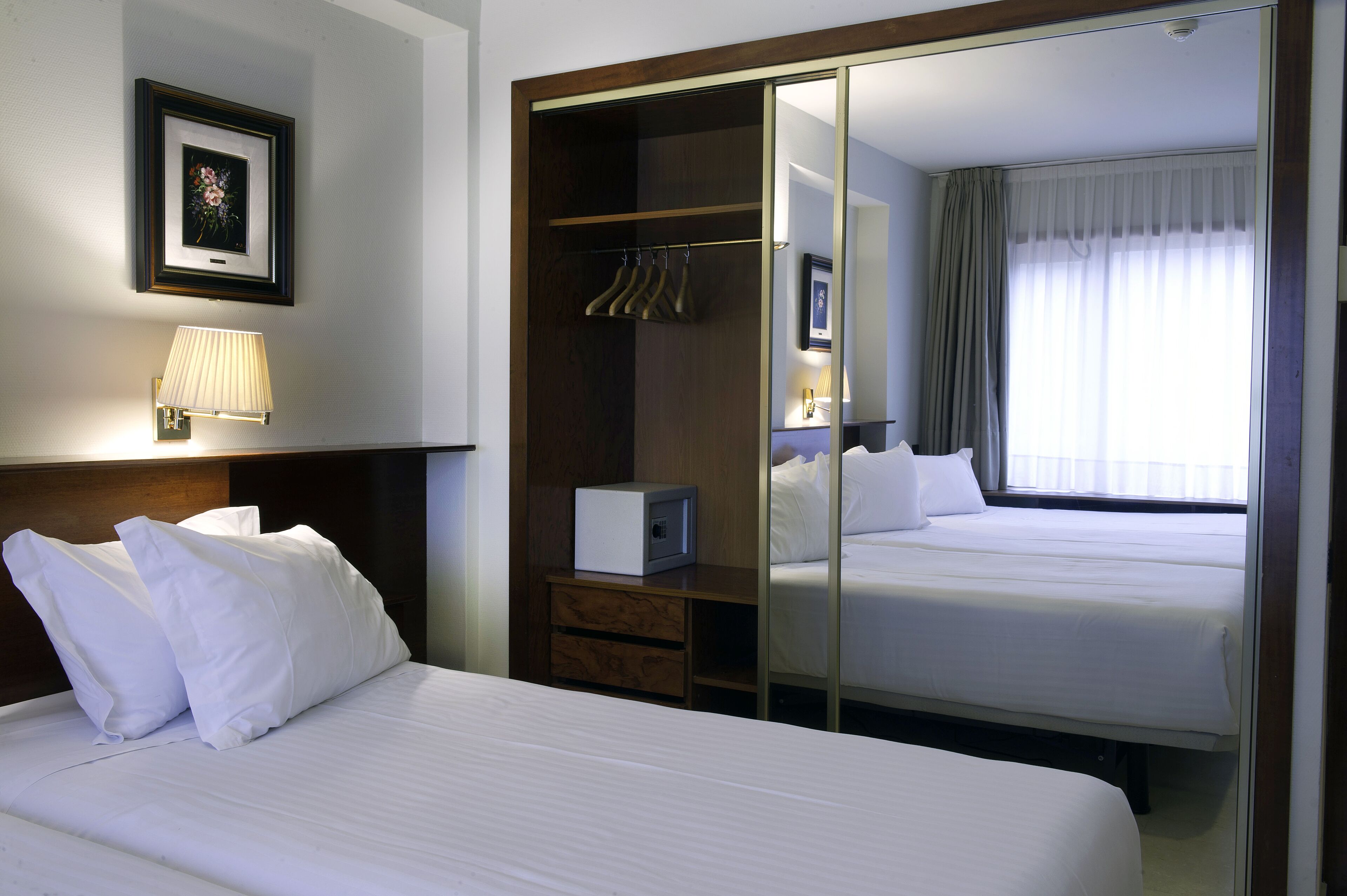 Habitación de hotel con una cama grande, un armario con ropa colgada, una mesita de noche con una lámpara y un espejo.