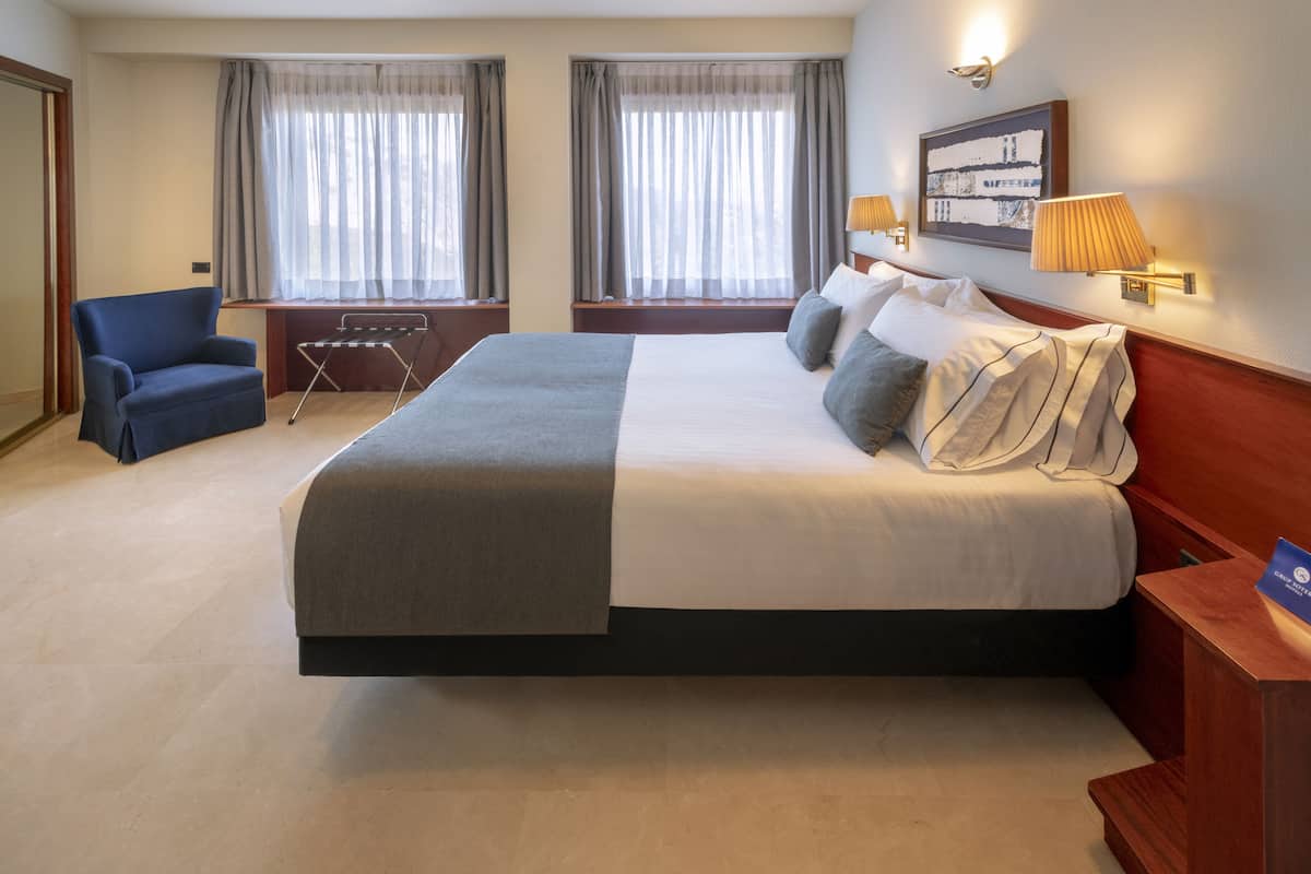 A hotel room with a large bed, two windows with curtains, a chair, a desk, and a wall-mounted lamp.