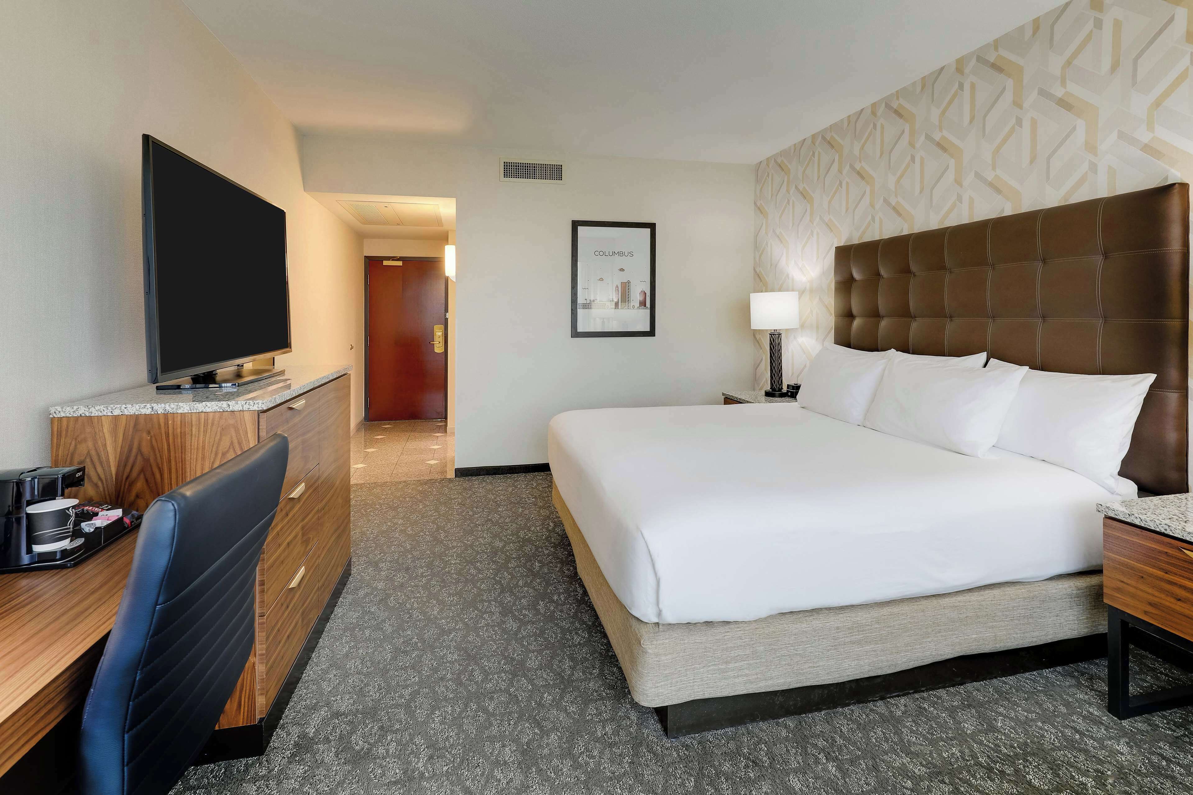 Een hotelkamer met een bed, een bureau met een televisie, een koffiezetapparaat, een hoofdeinde en een ingelijste foto aan de muur.
