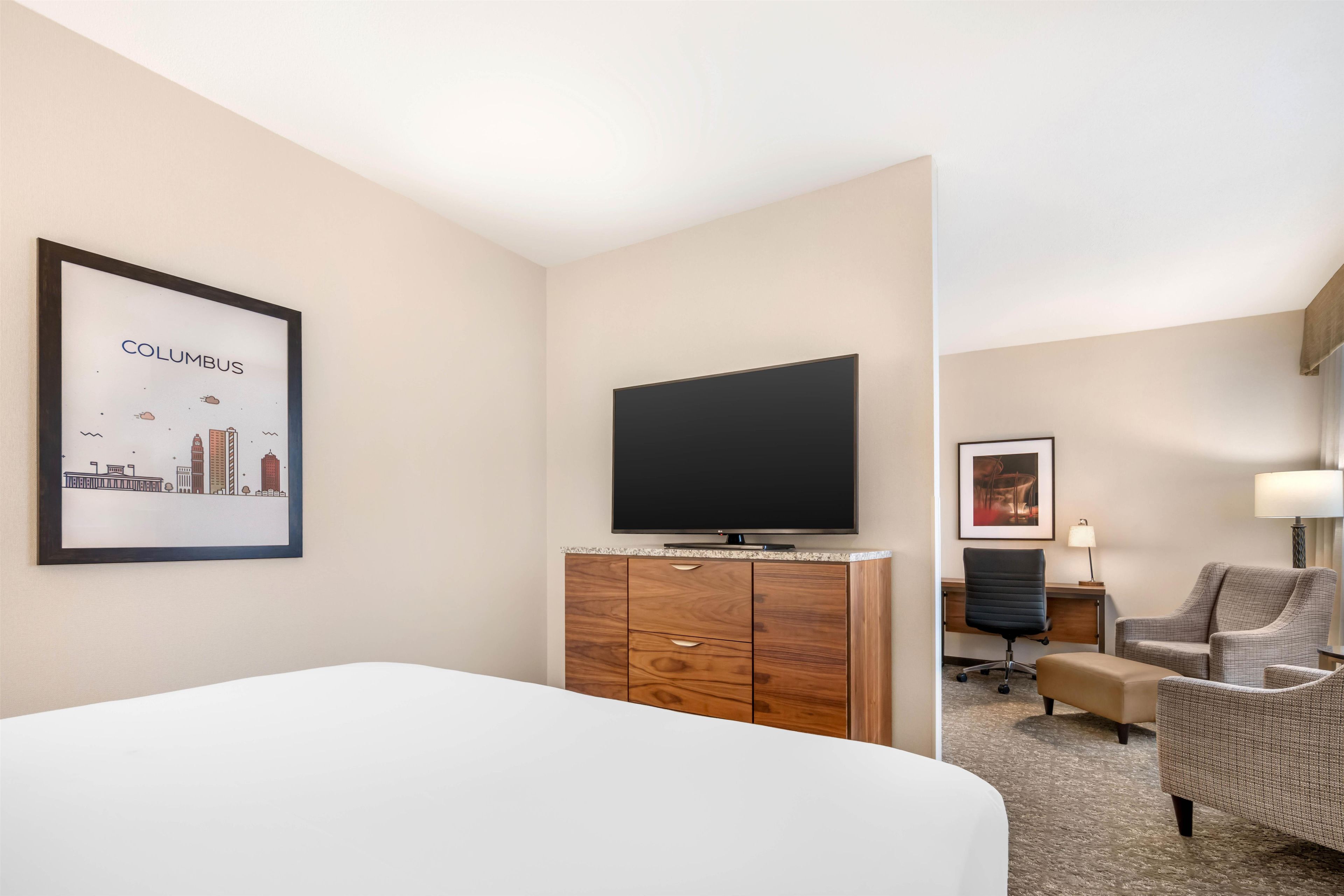 Quarto de hotel com cama, televisão de tela plana, cômoda de madeira, cadeira, escrivaninha e quadro na parede.