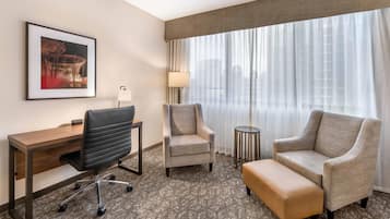 A hotel room with a desk, two armchairs, a small round table, a lamp, and a large window with curtains.