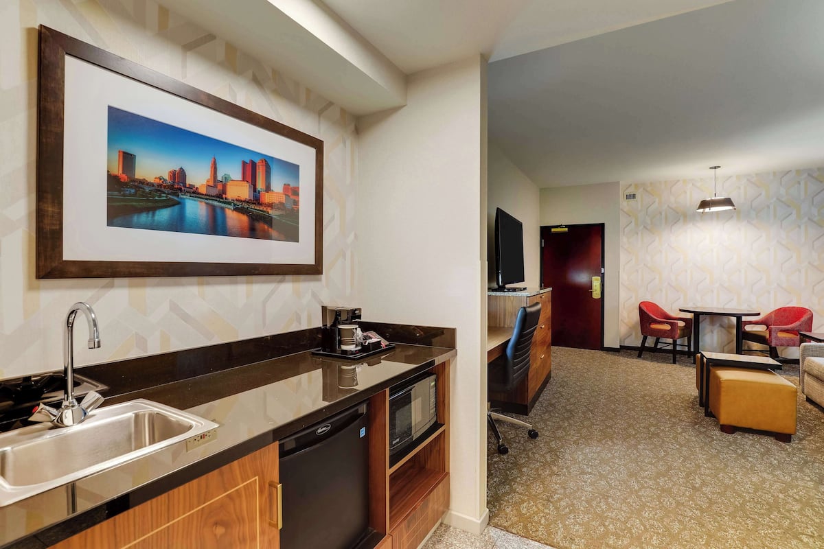 A compact hotel room with a kitchenette, a flat-screen TV, a dining area with red chairs, and a patterned carpet.