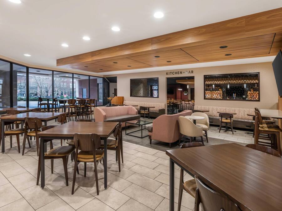 A modern restaurant with wooden tables and chairs, a kitchen bar area, and large windows offering views of the city.