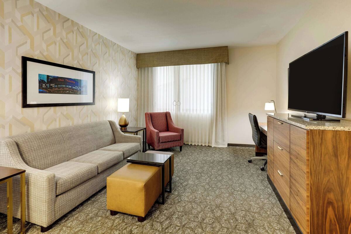 A hotel room with a sofa, armchair, ottoman, TV, and a framed picture on the wall.
