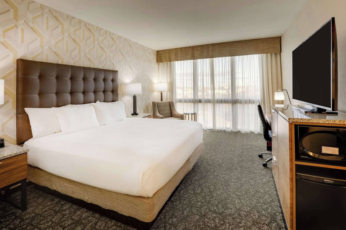 A hotel room with a large bed, two bedside tables, a desk with a flat-screen TV, and a view of the city through large windows.