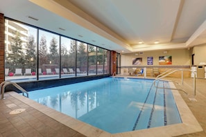 An indoor swimming pool with clear glass walls, a tiled deck, and lounge chairs outside.
