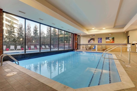 An indoor swimming pool with clear glass walls, a tiled deck, and a lounge area with chairs and a table.