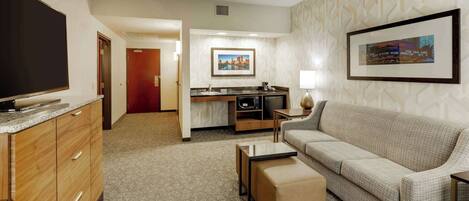 A hotel room with a flat-screen TV, a sofa, a coffee table, a kitchenette, and a framed picture on the wall.