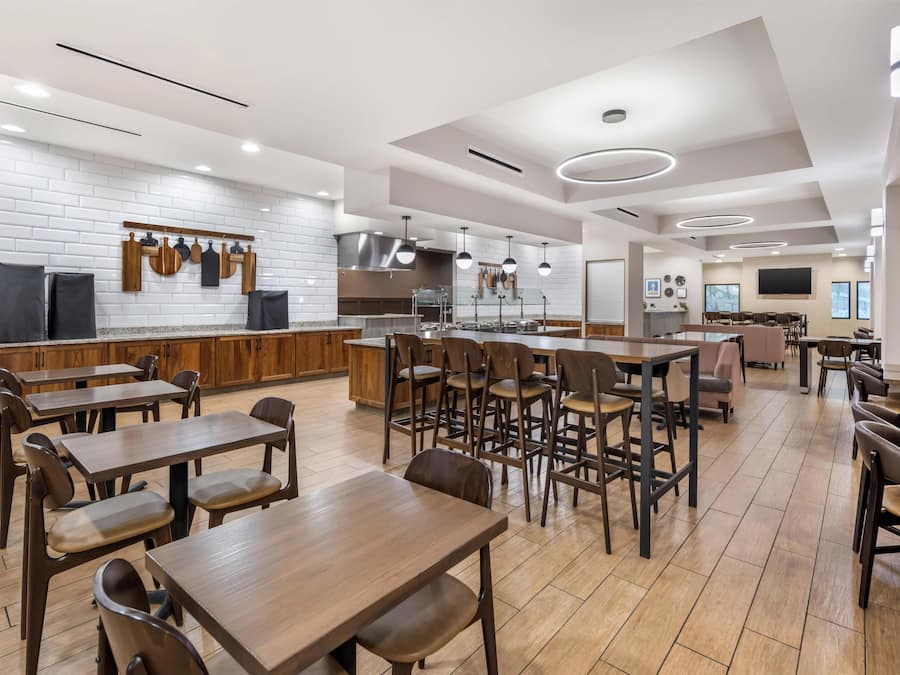 Un grand espace de salle à manger avec plusieurs tables et chaises, un coin cuisine équipé d’appareils électroménagers modernes et un téléviseur à écran plat fixé au mur.