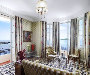 A luxurious room with a patterned floor, a chandelier, a painting, and a view of the sea. - Hotel Belles Rives (Antibes)