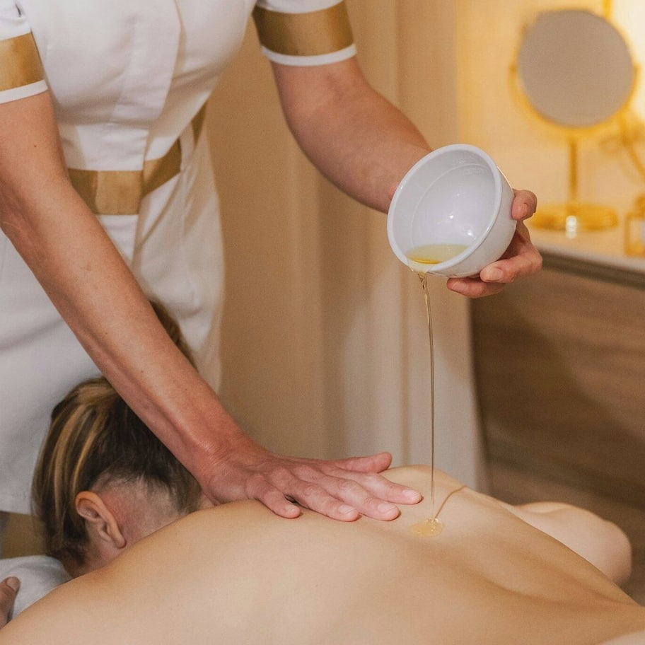 A person receiving a massage with a bowl of oil being poured onto their back.