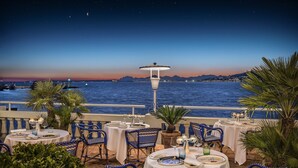 An outdoor dining area with tables and chairs, overlooking a body of water and a cityscape at dusk. - Hotel Belles Rives (Antibes)