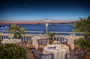 An outdoor dining area with tables and chairs, overlooking a body of water and a cityscape at dusk. - Hotel Belles Rives (Antibes)