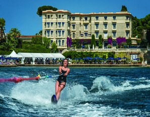 Une personne pratiquant le ski nautique sur une étendue d’eau, avec un bâtiment en arrière-plan.